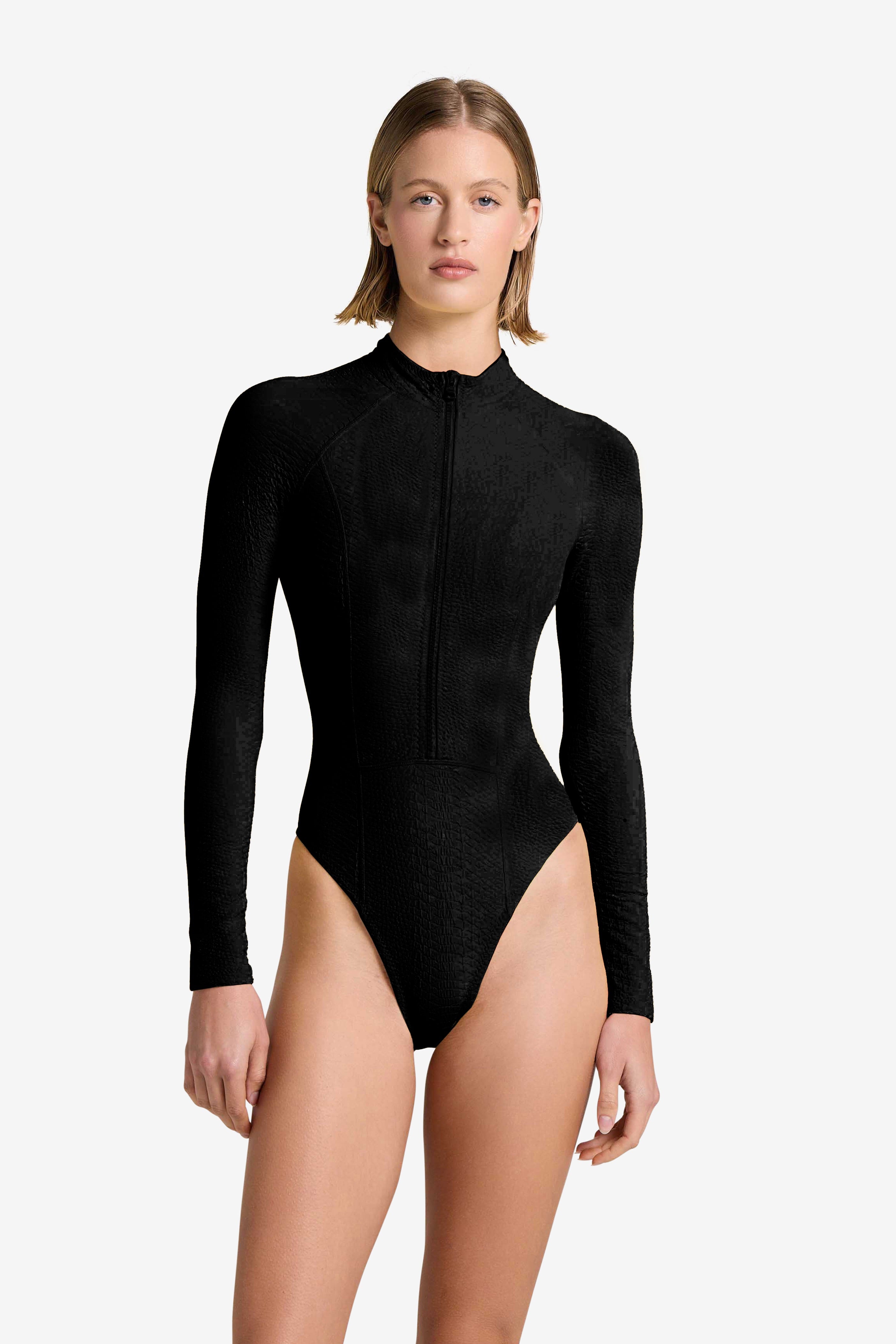 A person with short, straight hair wears the long-sleeve, black Sophie Snakeskin Surfsuit by Gigi C Bikinis, featuring a high neckline and snakeskin-embossed fabric—ideal for water sport enthusiasts—standing against a plain light background.