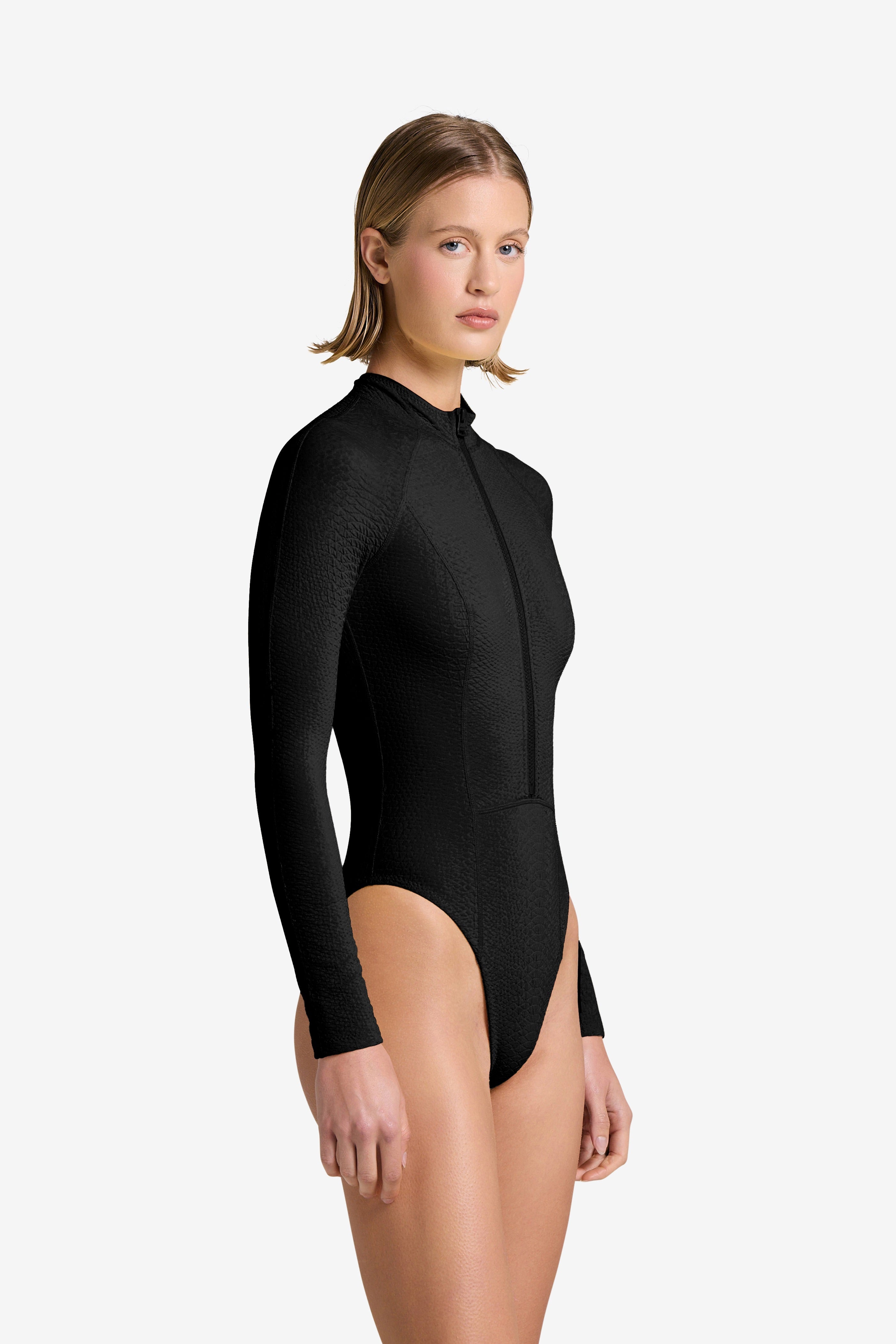 A woman with short, straight hair wears the fitted, long-sleeve black Sophie Snakeskin Surfsuit by Gigi C Bikinis, featuring a high neckline and front zipper in snakeskin-embossed fabric, standing against a plain light background.