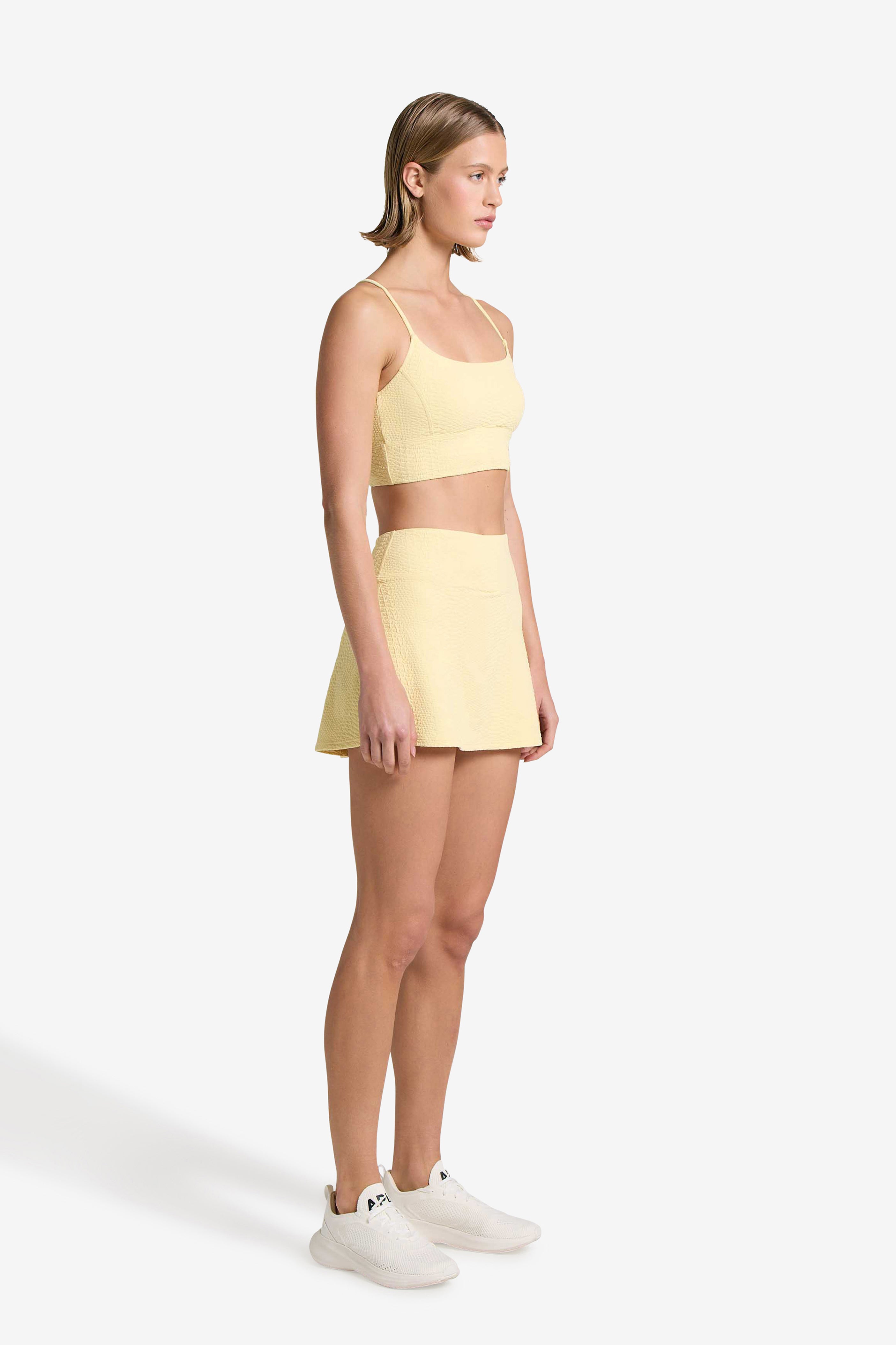 A woman wears a yellow sports bra and matching Gigi C Sport Heidi Snakeskin Skort with built-in shorts and white athletic shoes, standing against a white background and facing left with a neutral expression.