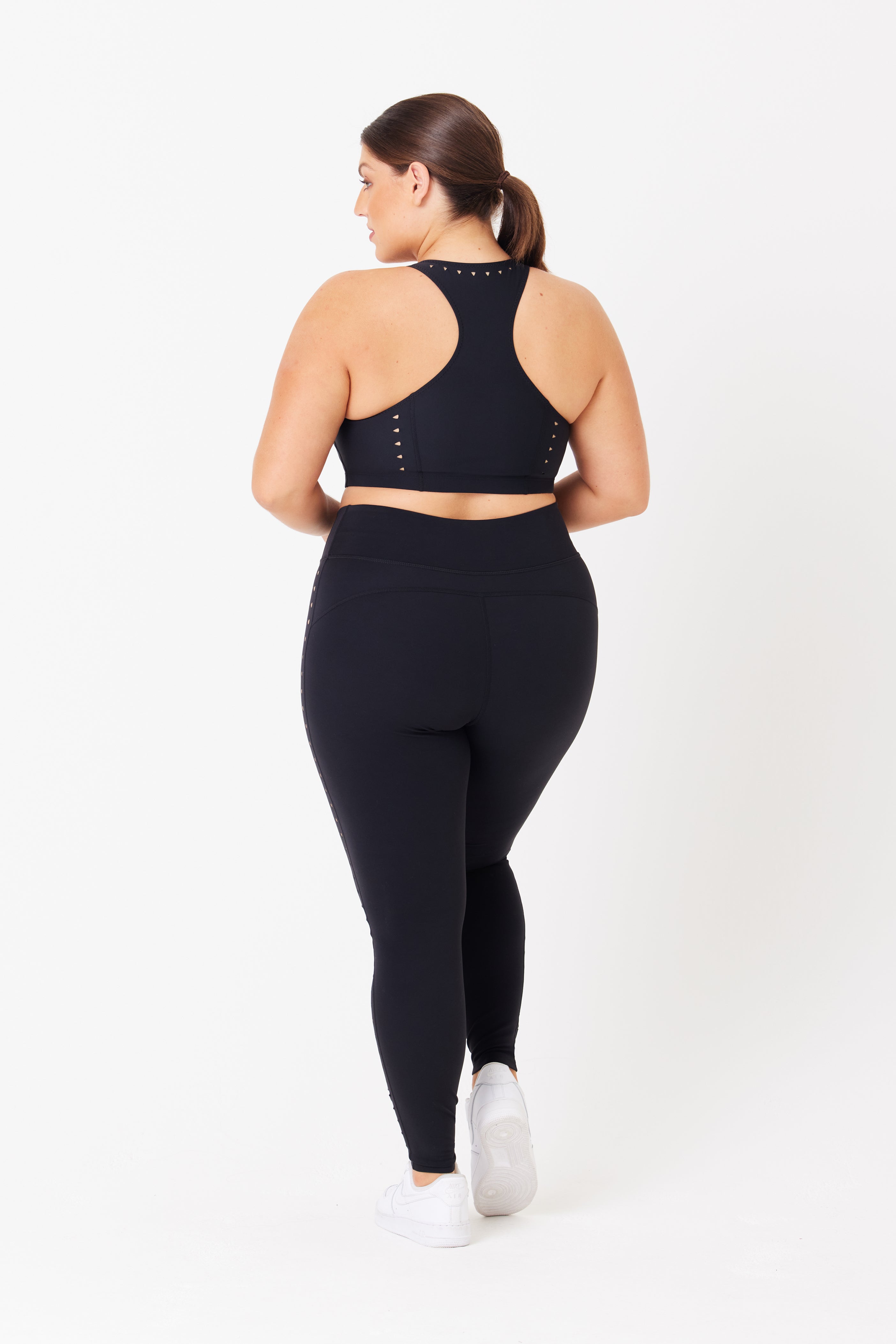 A person wearing a black sports bra with small laser cutouts and Katrina Legging by Gigi C Sport stands in white sneakers against a plain white background, facing away from the camera.