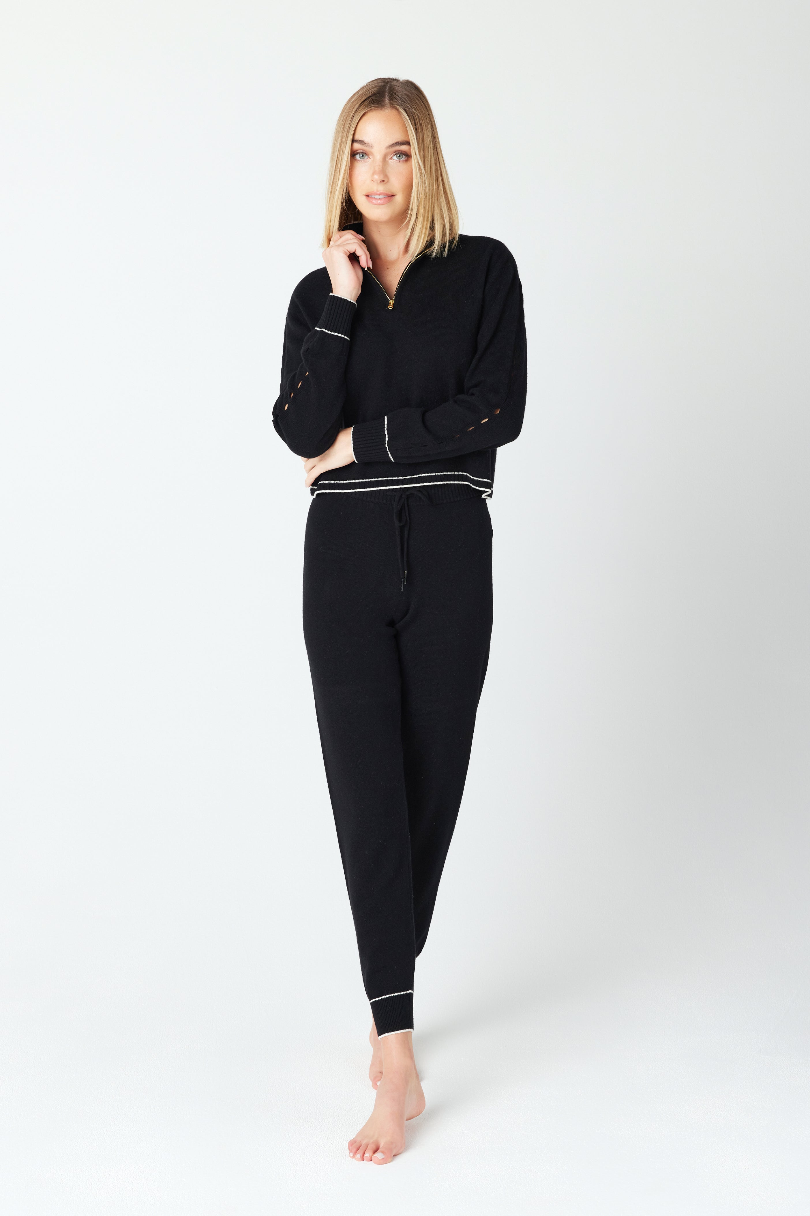 A person with shoulder-length blonde hair poses barefoot in Gigi C Lounges cozy, sustainable fashion—a black long-sleeve top and Alice Jogger made of a wool/cashmere blend with subtle white trim. They stand against a plain white background, embodying comfy loungewear.