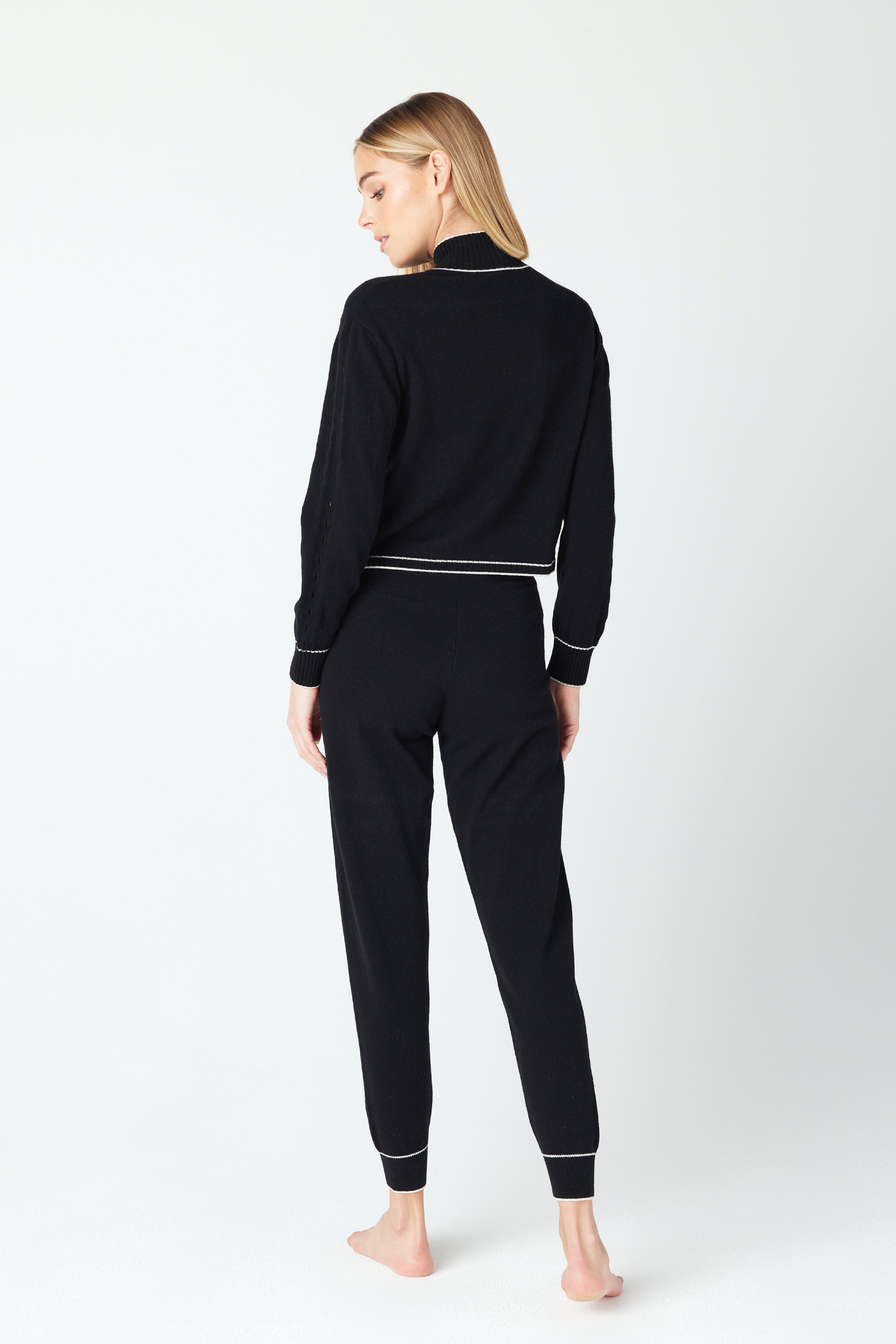 A person with long blonde hair stands barefoot, wearing the cozy Alice Jogger set by Gigi C Lounge—a sustainable black pajama ensemble with white trim, featuring a long-sleeve top and pants—radiating comfort against the light-colored background.