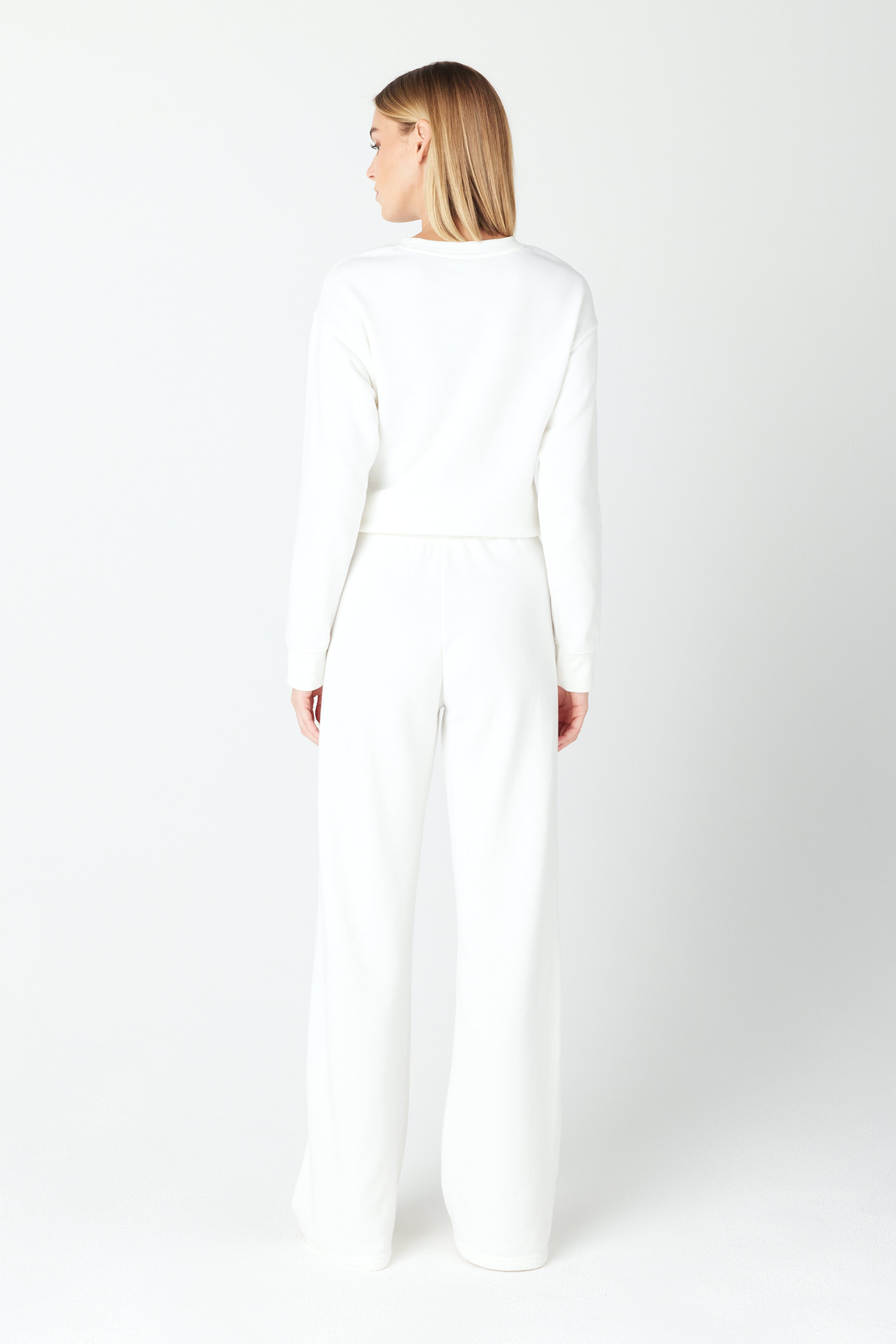 A person with long blonde hair stands facing away in a Kenzie Pullover by Gigi C Lounge and wide-legged pants. The Loop Terry Fleece texture contrasts the plain white background for a minimalist look.