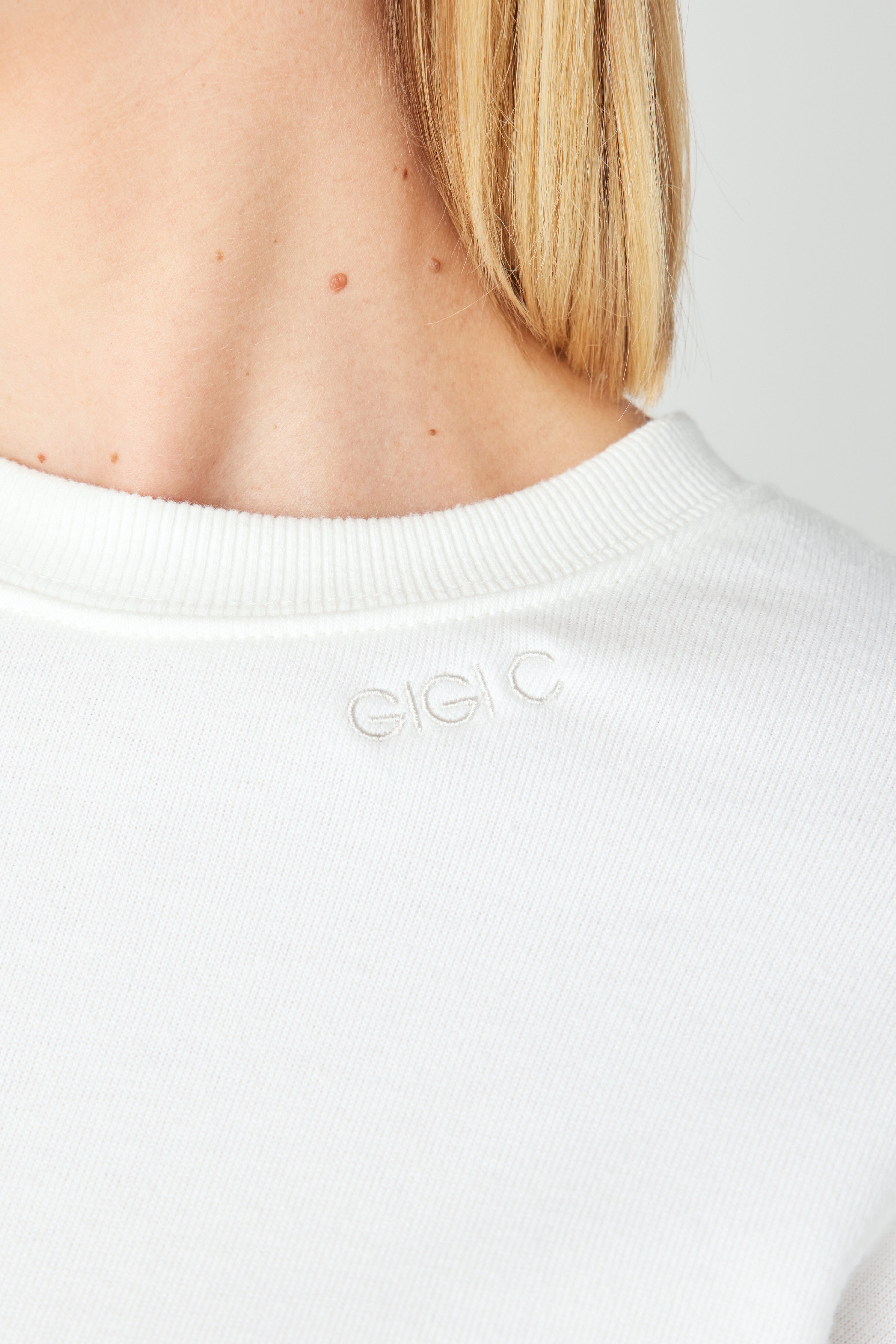 A close-up shows a person in a Kenzie Pullover by Gigi C Lounge, featuring small embroidery and crafted from soft Loop Terry Fleece. The background is plain and neutral.