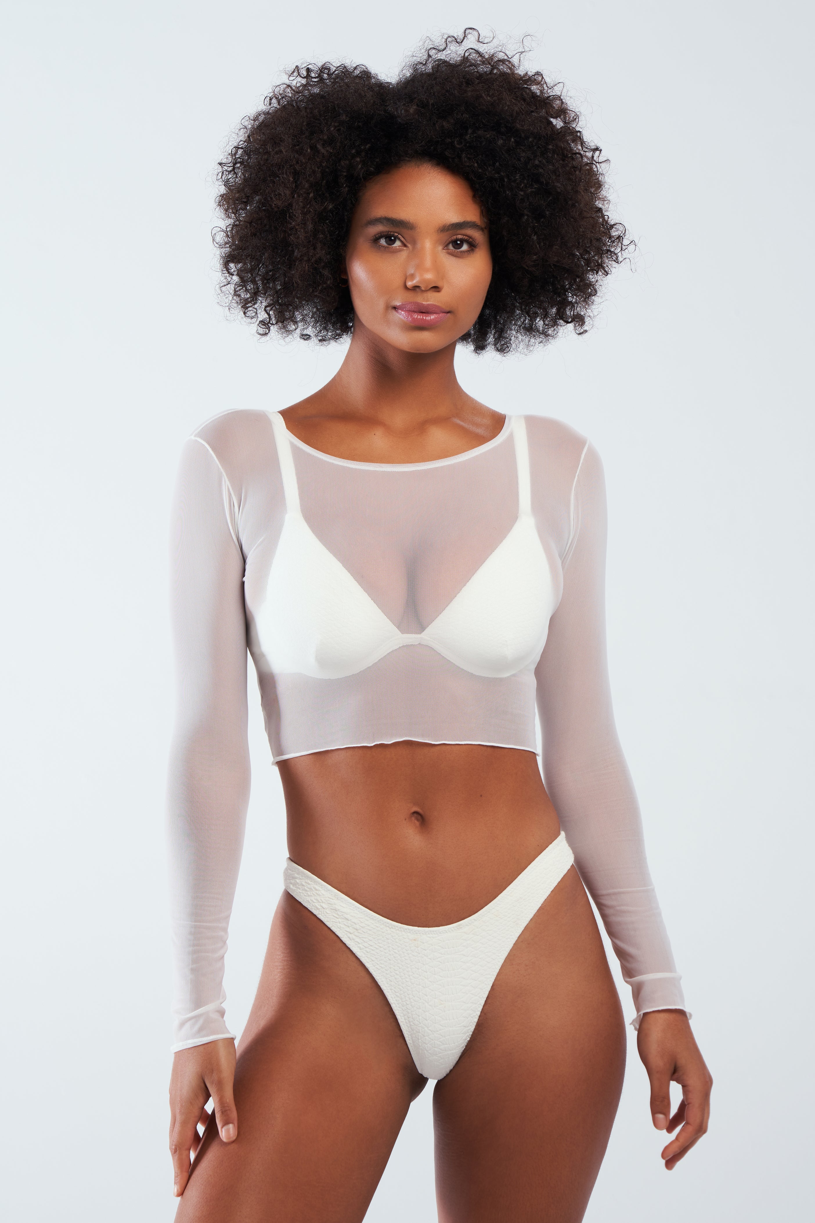 A person with curly hair dons the Gigi C Bikinis Caroline Mesh Crop Top over a white bra and underwear, creating a striking silhouette. They stand against a plain white background, highlighting the delicate design of their outfit.