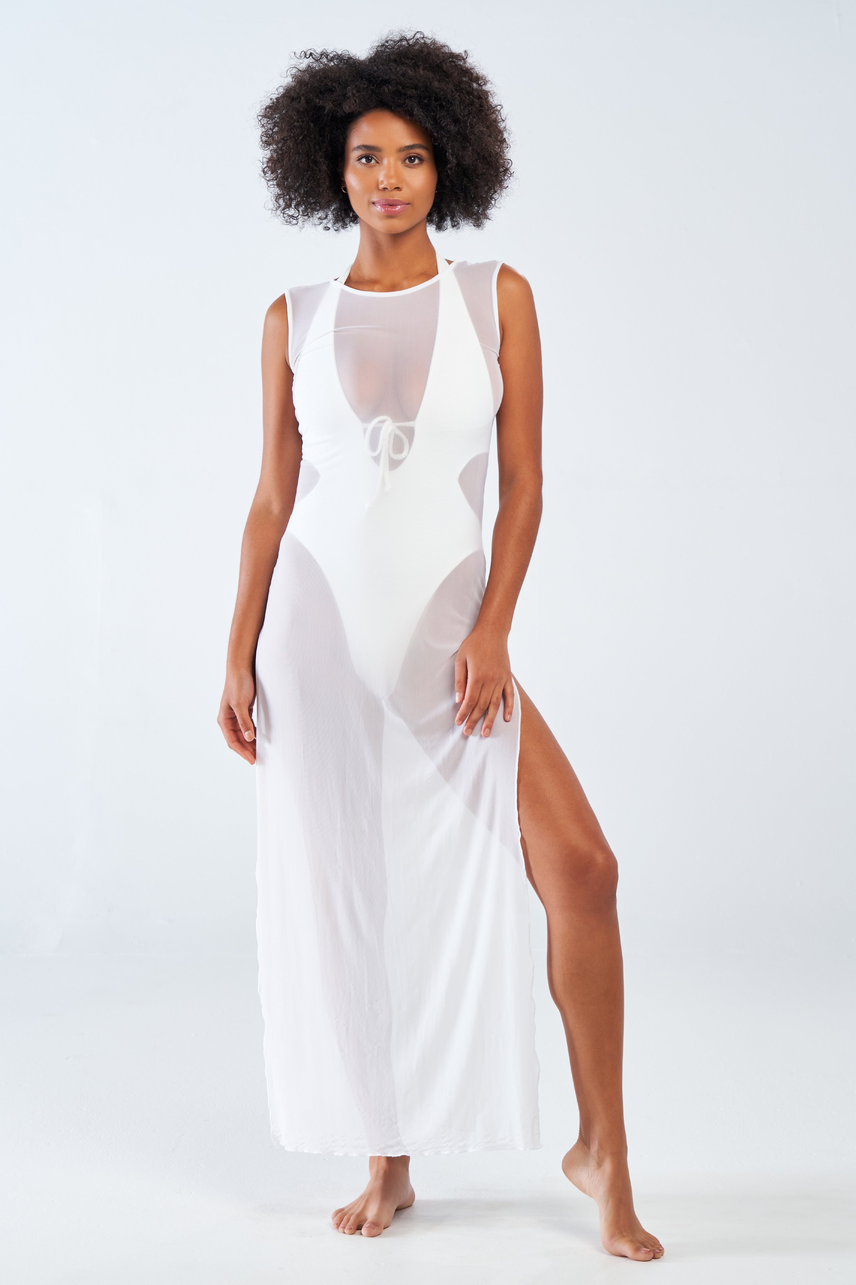 A person with curly hair is wearing the silky soft Terri Mesh Coverup from Gigi C Bikinis, featuring a sheer sleeveless design with a high slit and tie detail at the front. They stand barefoot against a plain white background, looking directly at the camera.