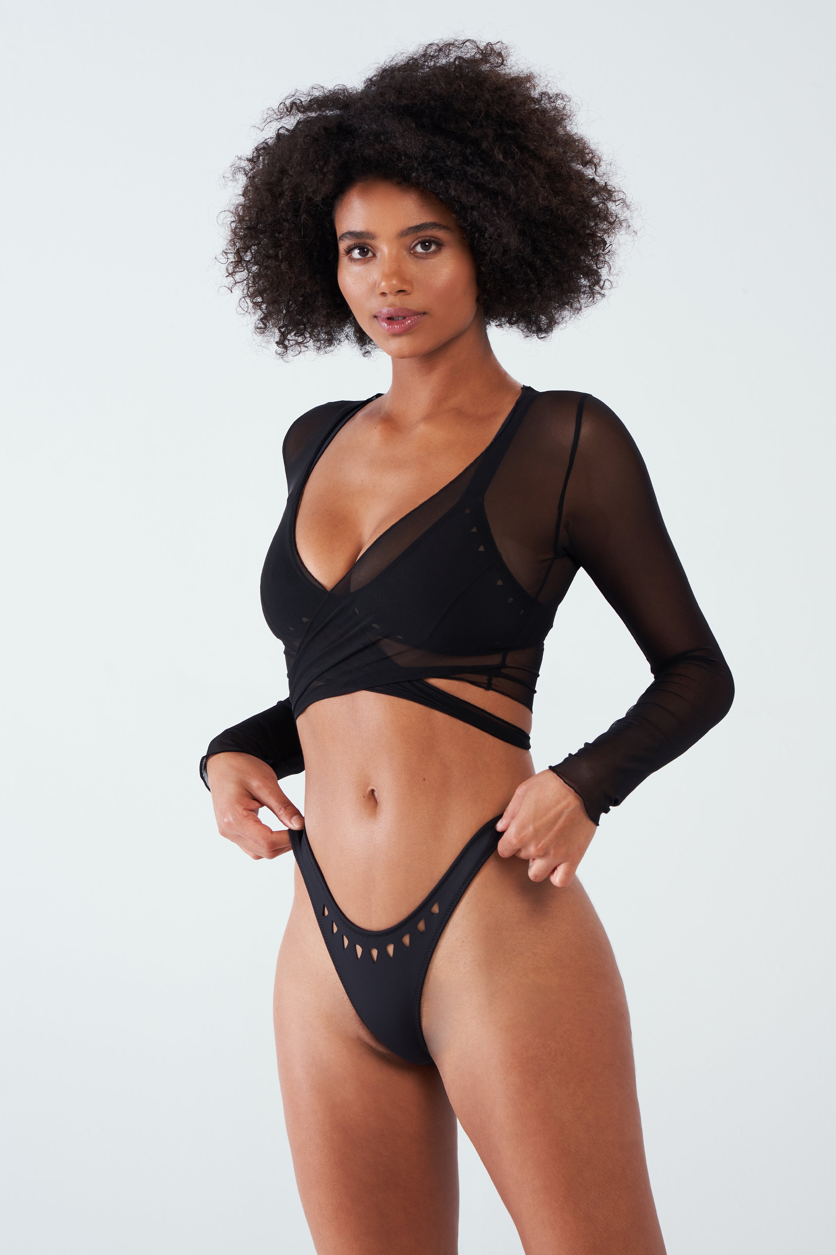 A person with curly hair confidently poses in a Gigi C Bikinis Tina Mesh Wrap Top, featuring sheer fine mesh and adjustable waist ties, paired with high-cut bottoms with cut-out details against a plain light background.