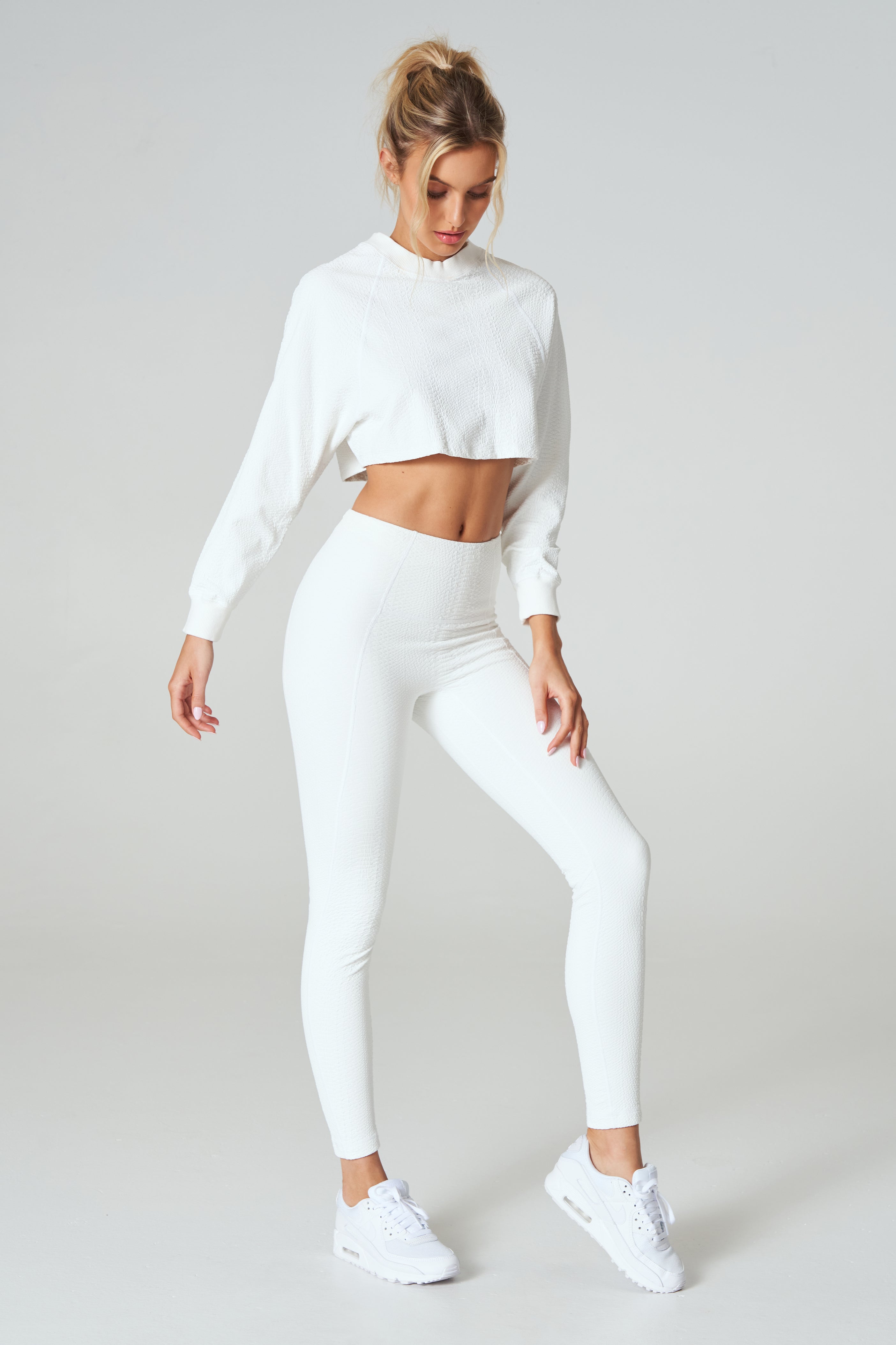 A person poses against a plain background wearing the Isabella Pullover by Gigi C Sport with matching leggings and white sneakers. They stand with one leg slightly bent, gaze downward, and have their hair tied up.
