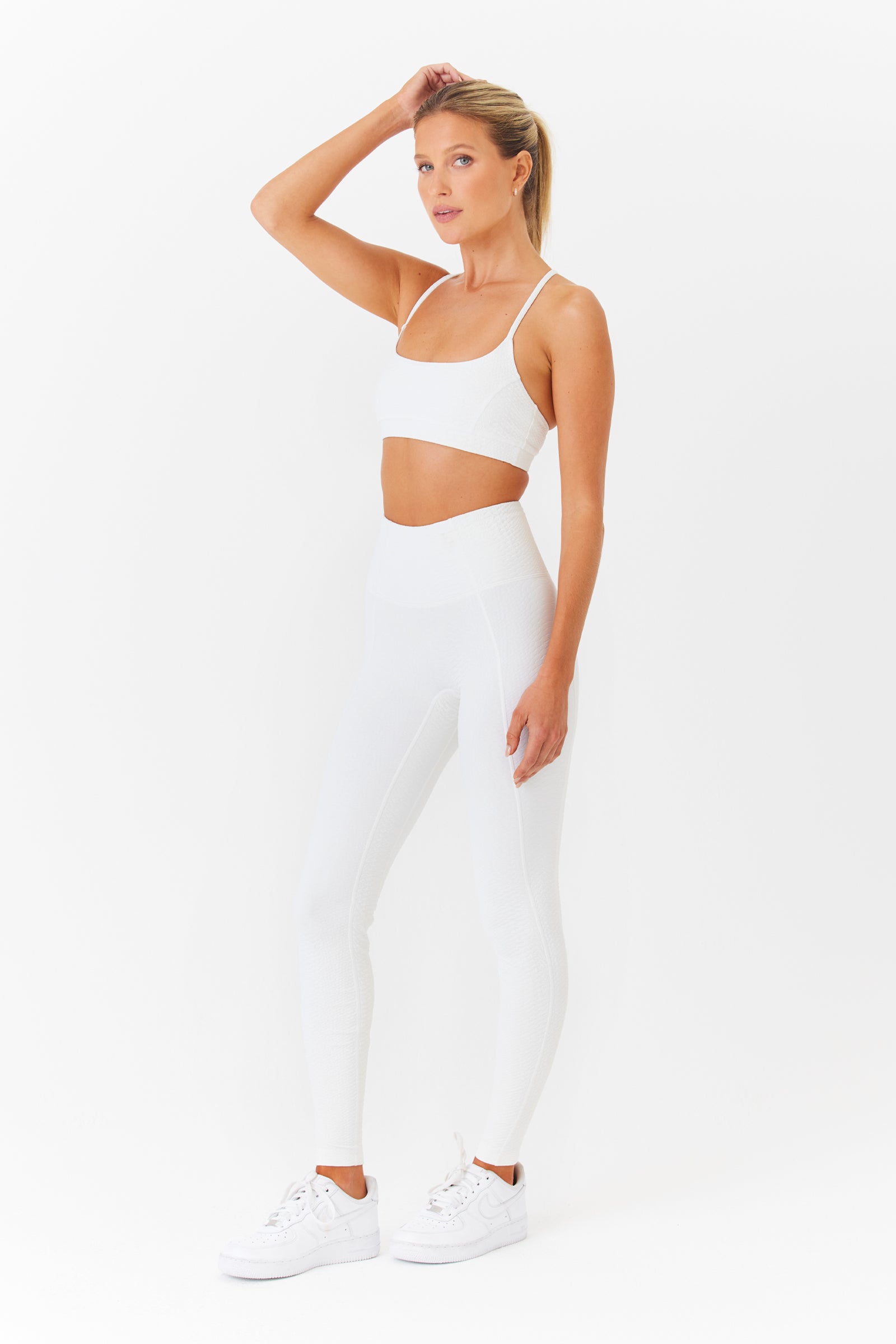 A woman poses in an Ava Legging by Gigi C Sport, paired with a white sports bra. Her right hand gently touches her hair as she stands in white sneakers against a plain backdrop. The bi-stretch compression fabric accentuates her movements while her left arm rests by her side.