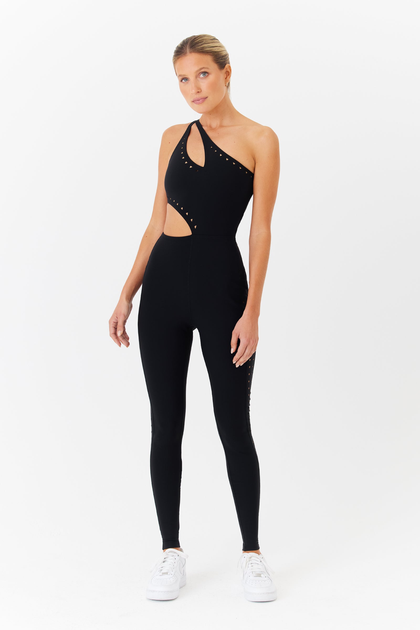 A person models the Gigi C Sports Cassie Jumpsuit, showcasing sleek laser cutouts, paired with white sneakers. The breathable knit design offers comfort and style as they pose against a plain white background.