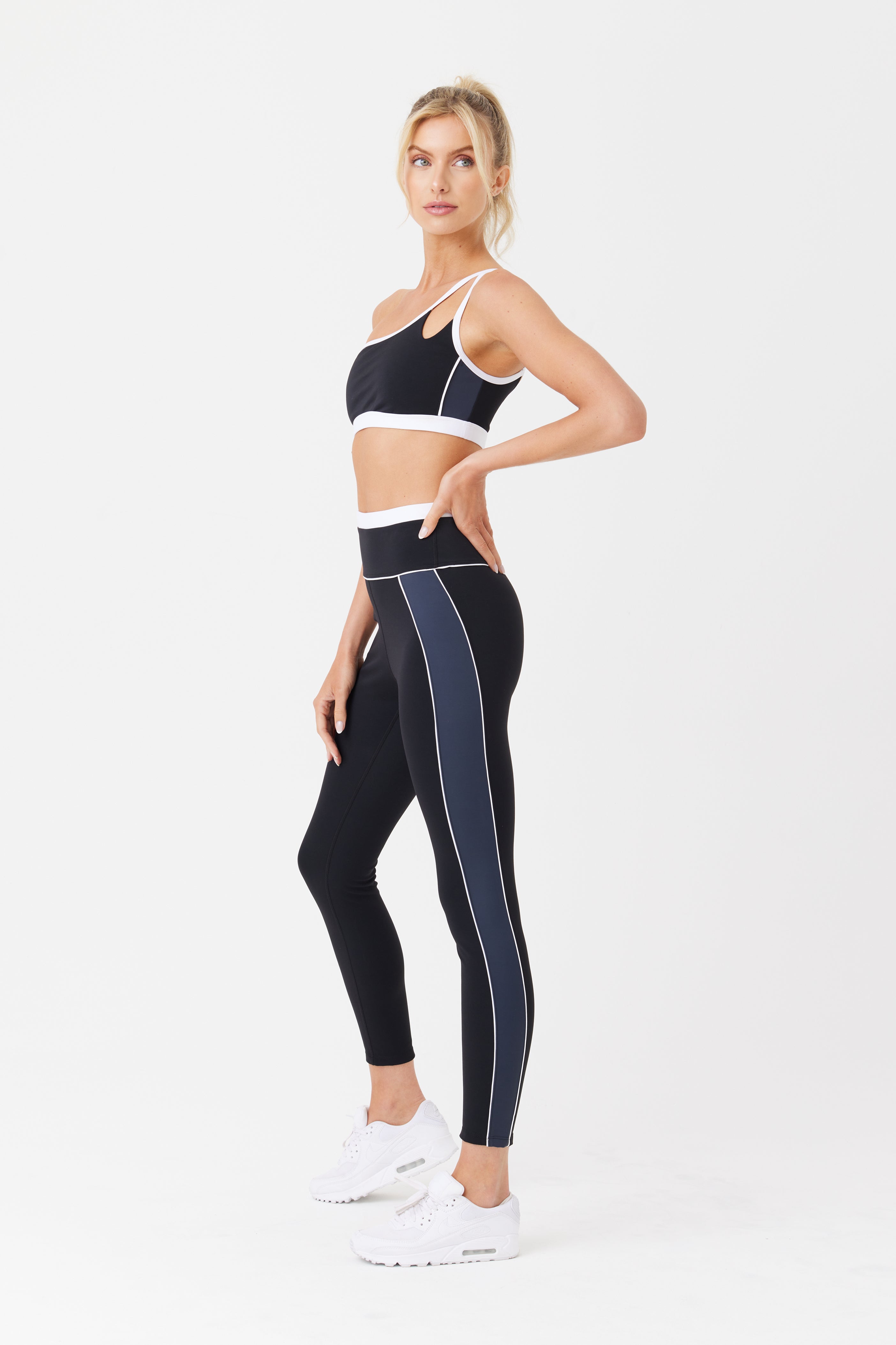 A woman models her Faith Asymmetrical Bra from Gigi C Sport, wearing black athletic wear with white trim. Paired with a crop top, high-waisted leggings, and white sneakers, she stands against a plain white background with her hair tied back.