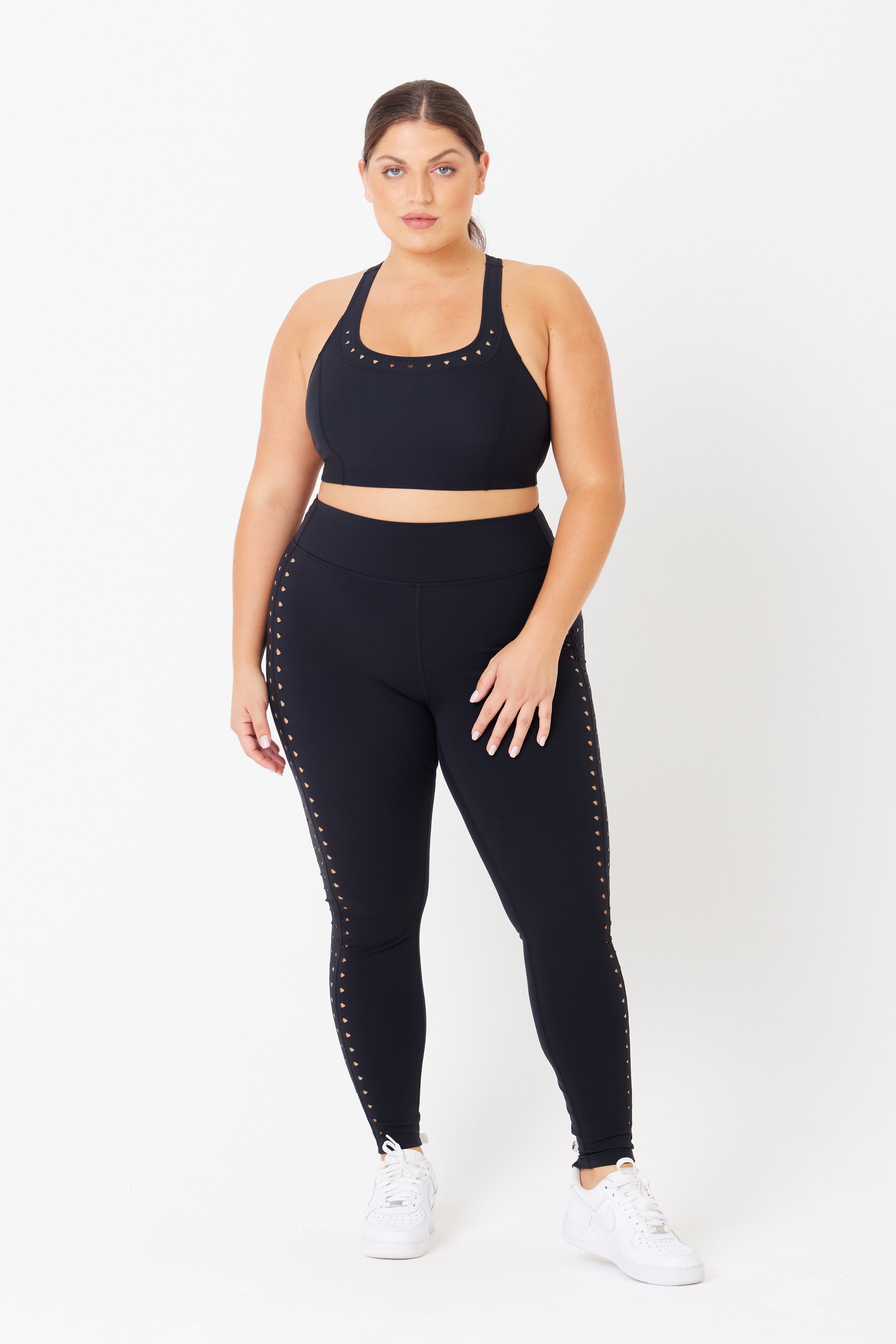 Against a white background, someone models the Annabelle Bra Top from Gigi C Sport and matching leggings with stud detailing. Made from breathable knit and recycled materials, they complement it with white sneakers and tied-back hair.