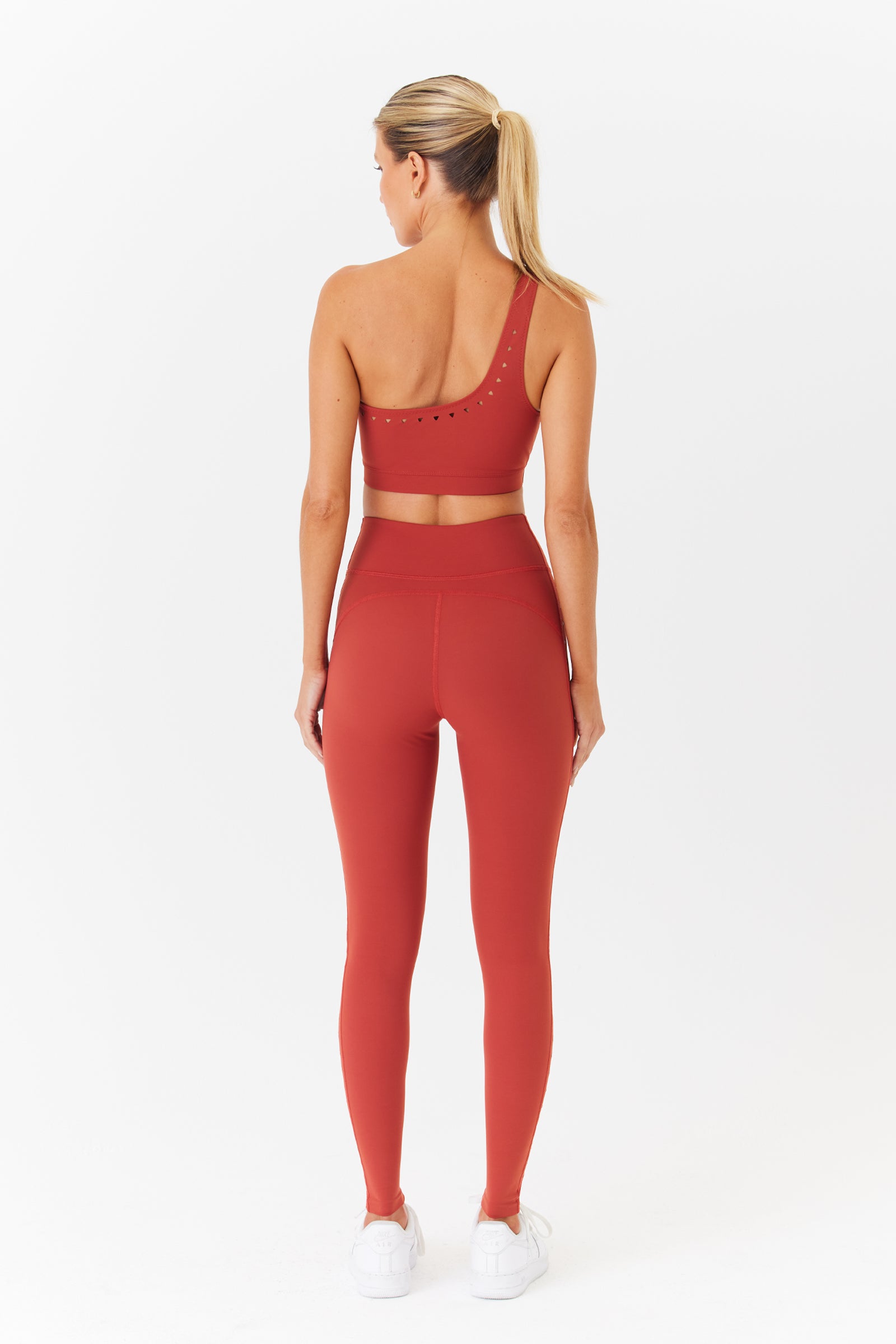 With a ponytail and back to the camera, a person is ready for hot yoga in a Jacqui Asymmetrical Bra by Gigi C Sport, high-waisted leggings, and white sneakers against a plain white background.