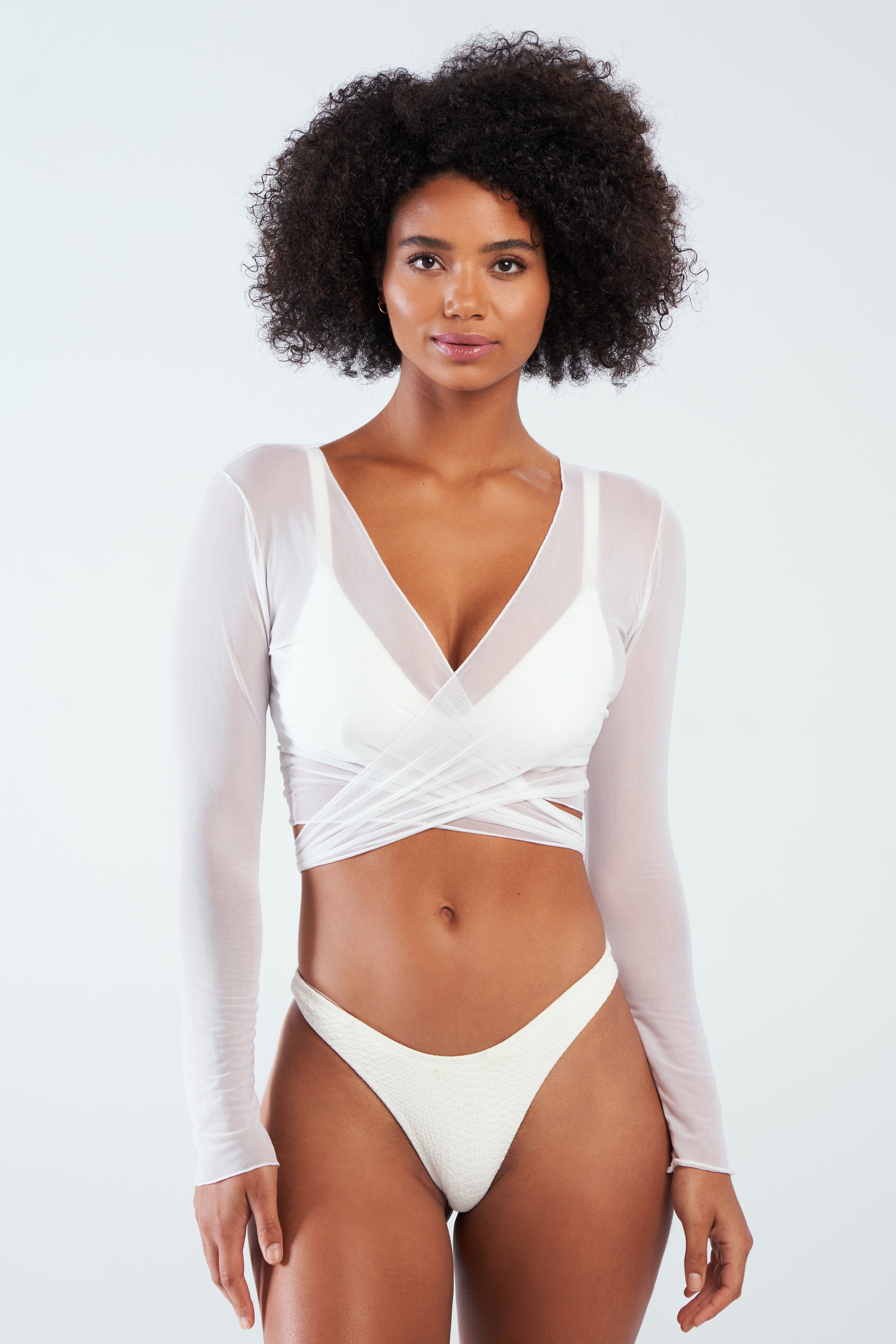 A woman with curly hair models the Gigi C Bikinis Tina Mesh Wrap Top featuring a plunging neckline, paired with white bikini bottoms, standing against a plain white background with a neutral expression.