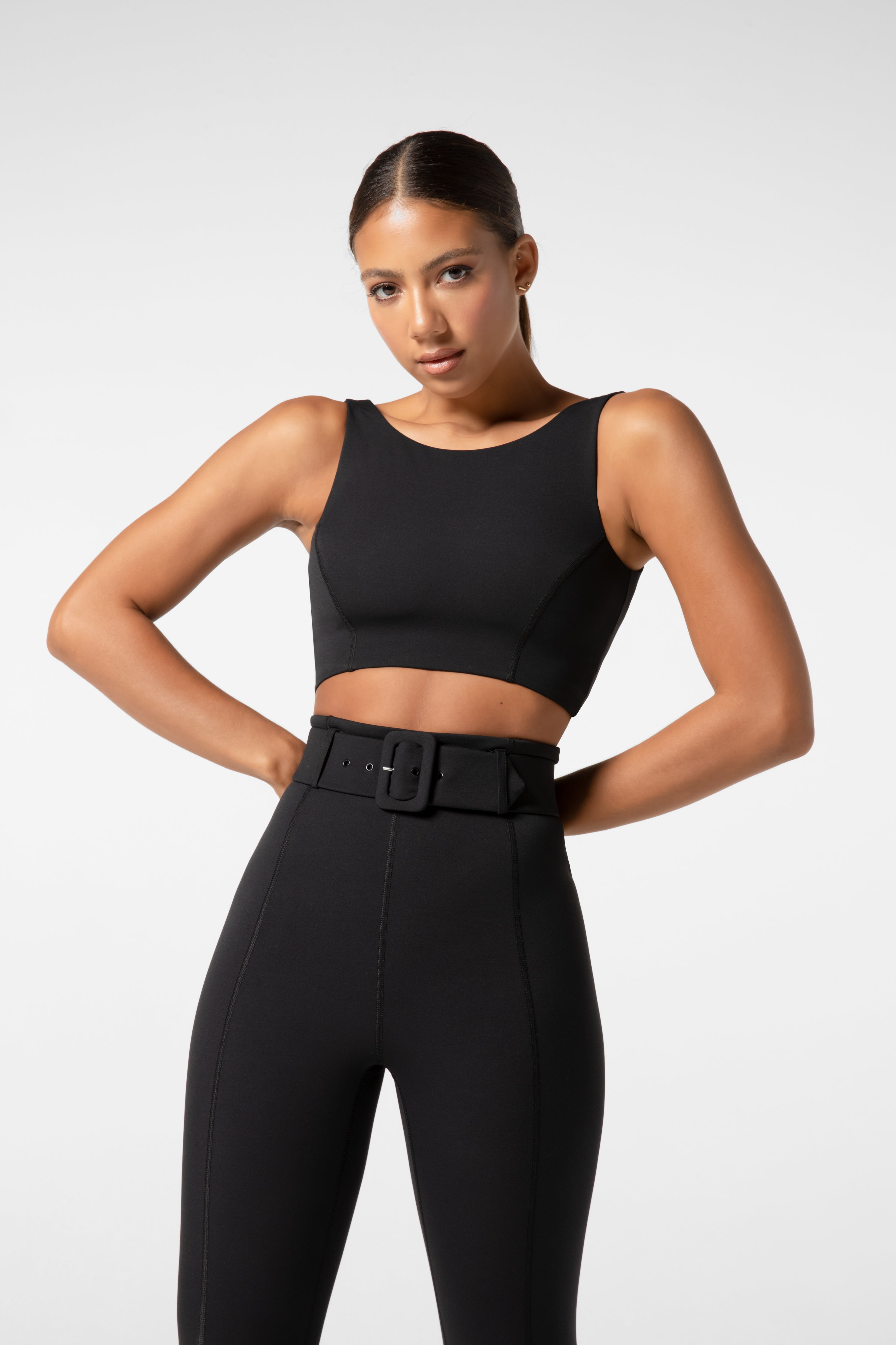 Someone wearing a Lola Tank Top by Gigi C Sport and high-waisted pants with a belt stands confidently against a plain background. Their hair is pulled back, hands on hips, the removable bust pads and breathable fabric ensuring comfort and coolness.